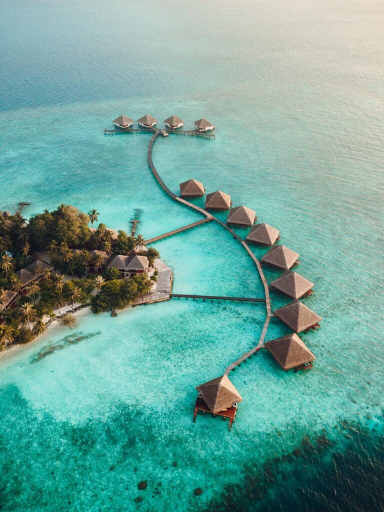 Over-water bungalows with thatched roofs connected by walkways in turquoise tropical waters.