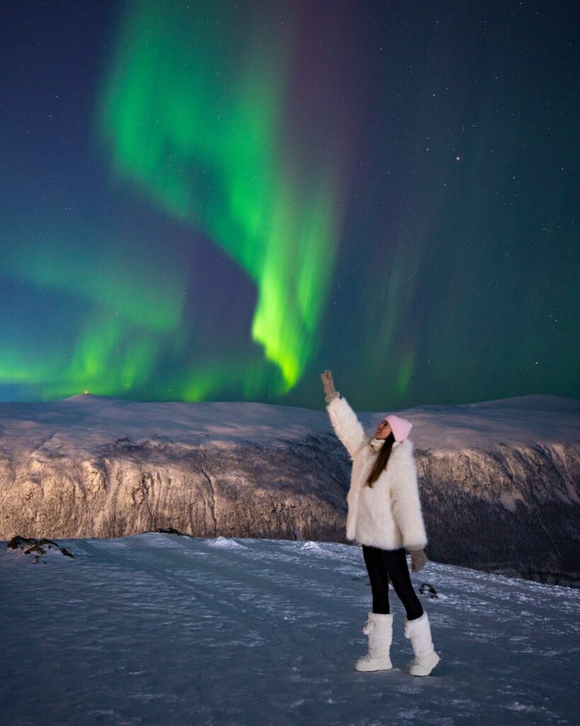 Woman in white winter gear pointing at green aurora borealis over snowy mountains