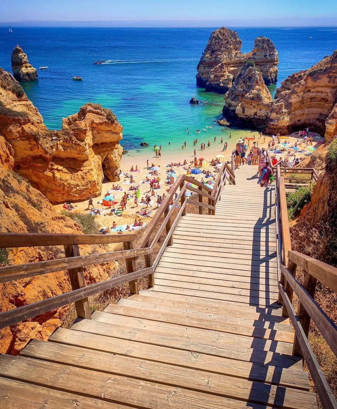 Wooden stairs leading down to a sunny beach with turquoise water and cliffs.