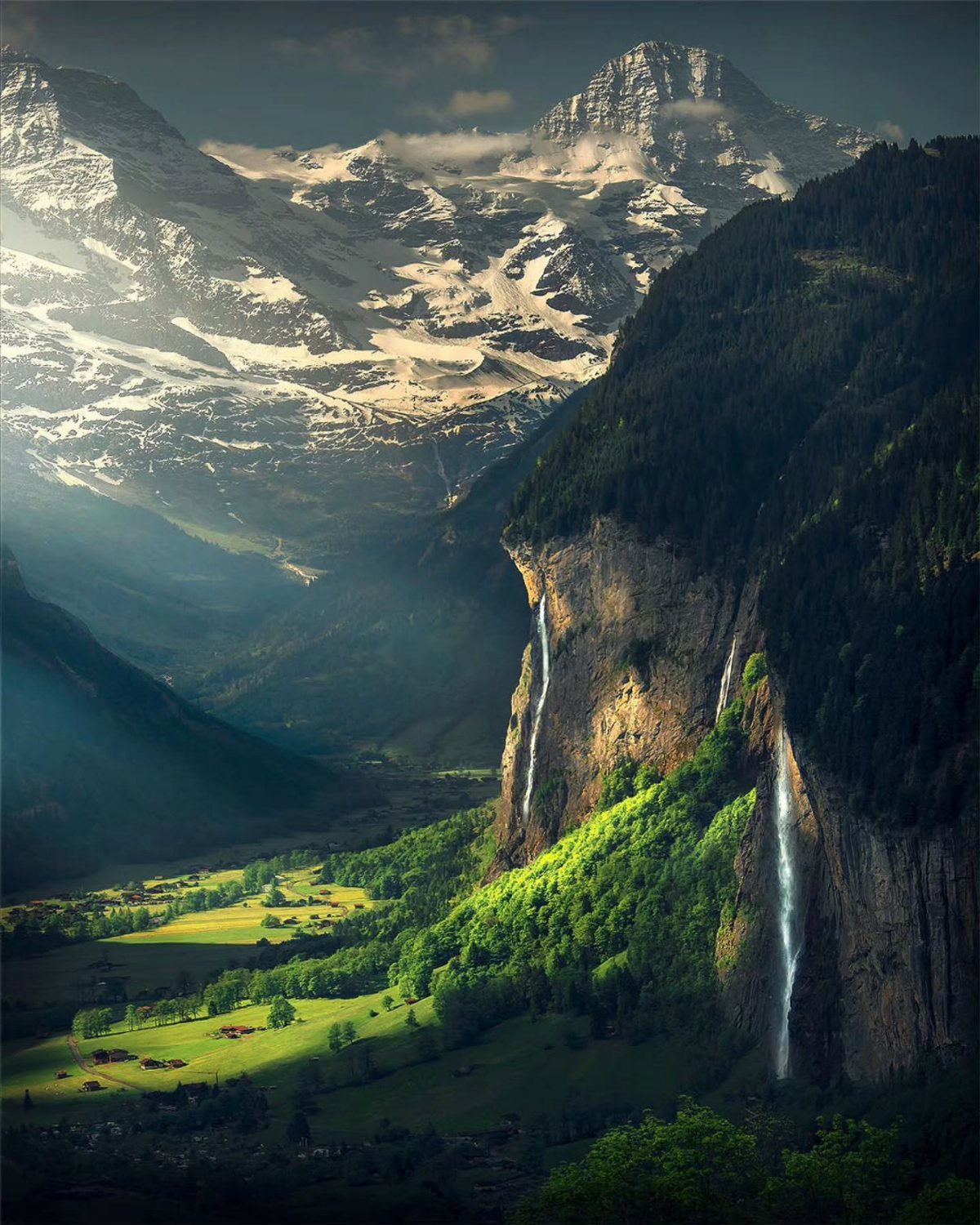 Snowy mountain peaks overlooking a green valley with waterfalls falling from cliffs.