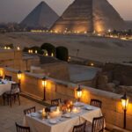 Dinner table on a terrace overlooking the illuminated Pyramids of Giza at dusk.