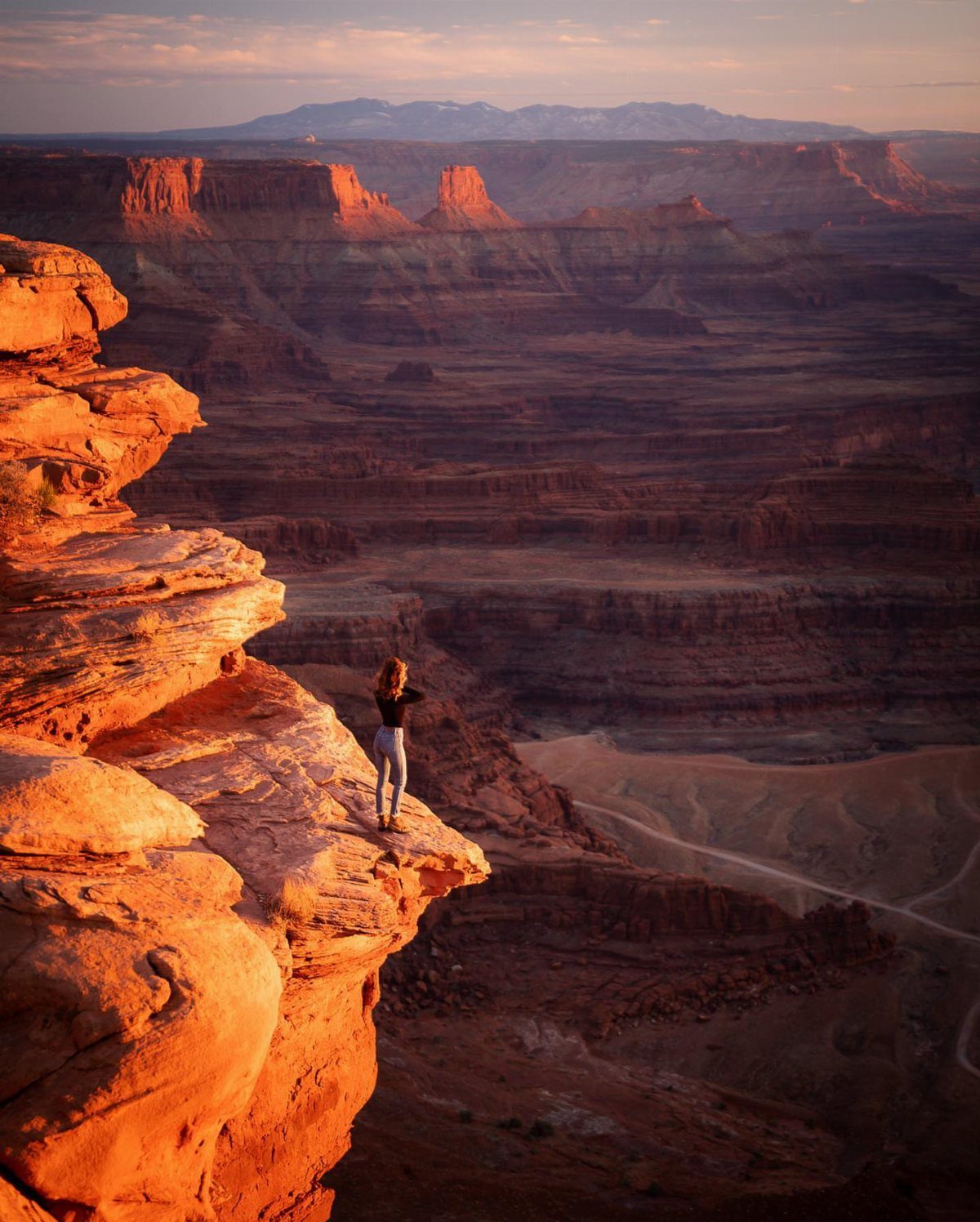 Person standing on a red rock cliff overlooking a vast desert canyon landscape.