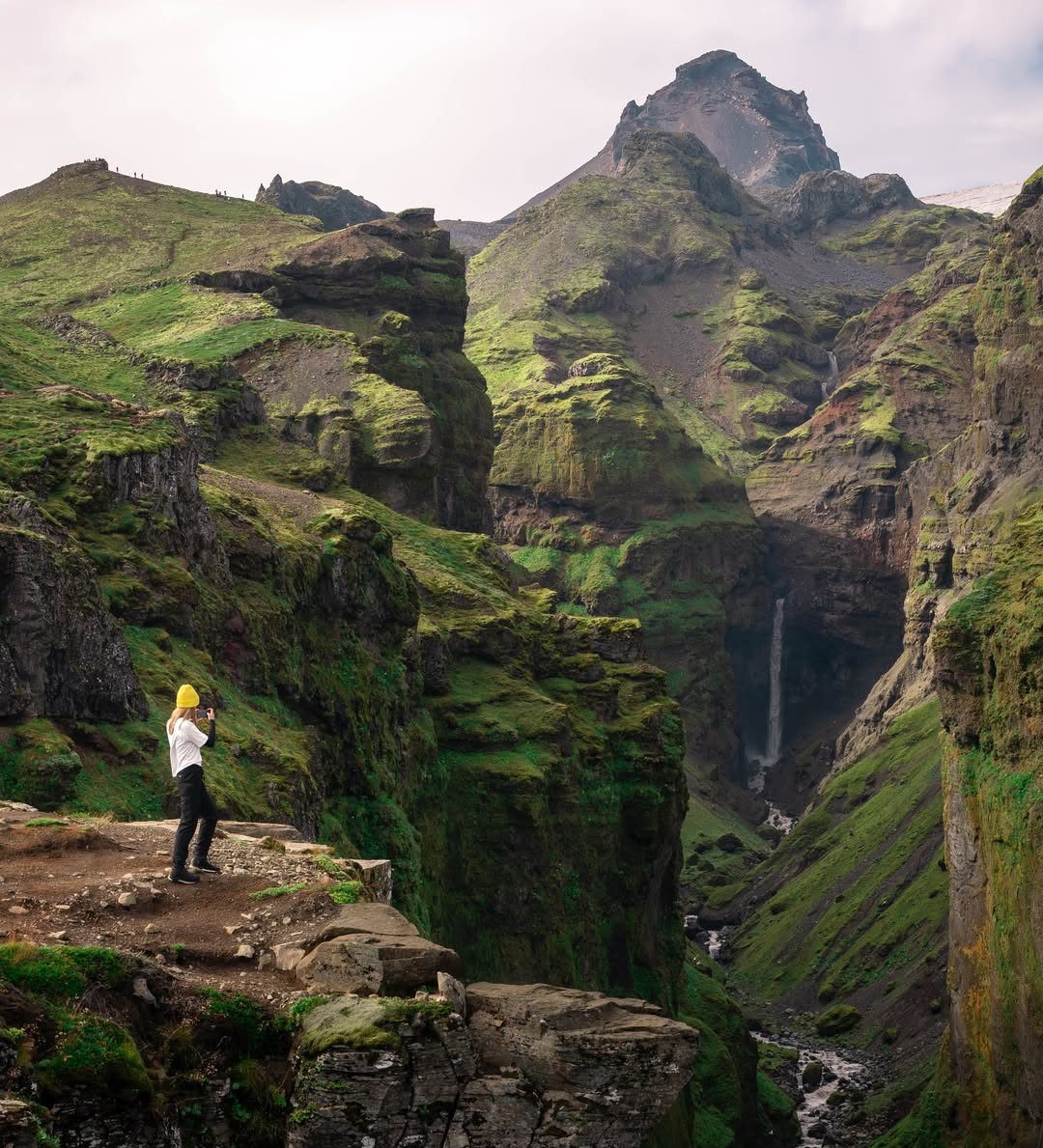 Person standing on a cliff overlooking a lush green valley with a waterfall.