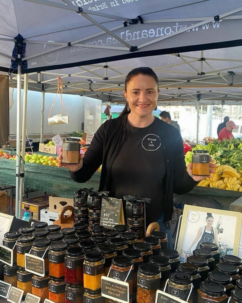 Woman holding jars of preserves at a local market stall with fresh produce.