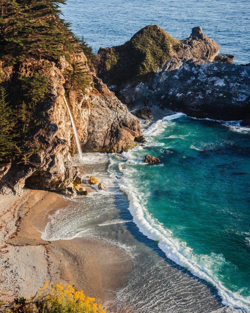 Waterfall cascading down a rocky cliff onto a sandy beach and turquoise ocean.