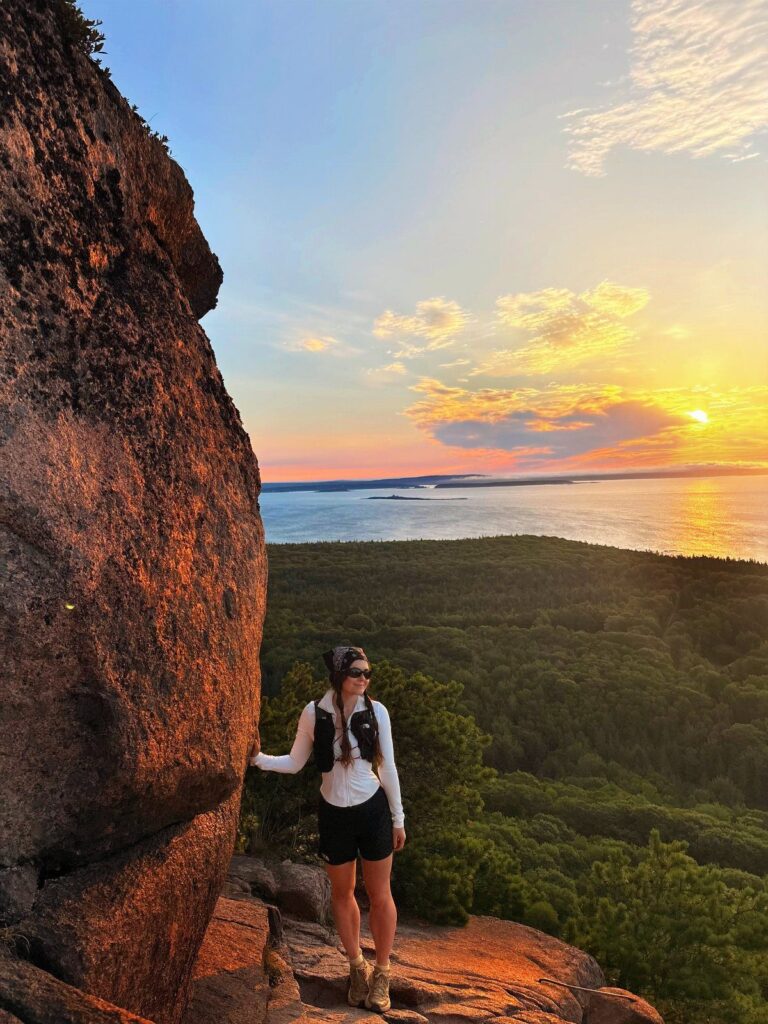 Woman standing on a rocky cliff overlooking a forest and ocean at sunset.