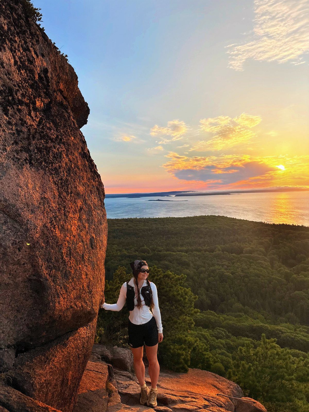 Woman standing on a rocky cliff overlooking a forest and ocean at sunset.
