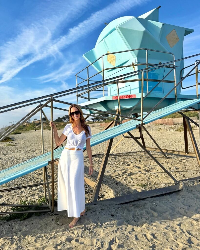 Woman in white clothing posing by a blue geometric lifeguard tower on a beach