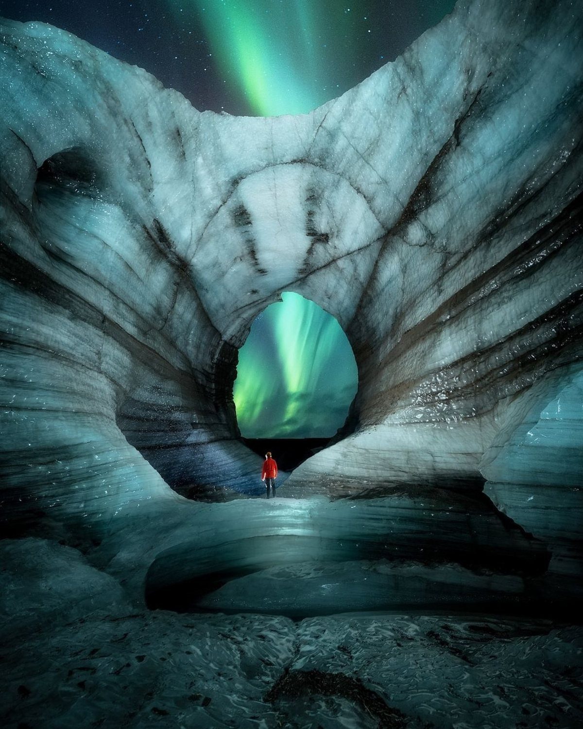 Person in red jacket inside an ice cave viewing green northern lights