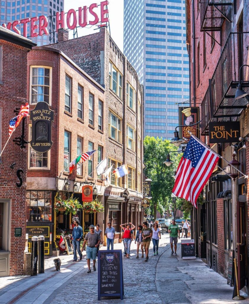 Cobblestone street in Boston with red brick buildings, American flags, and pedestrians.