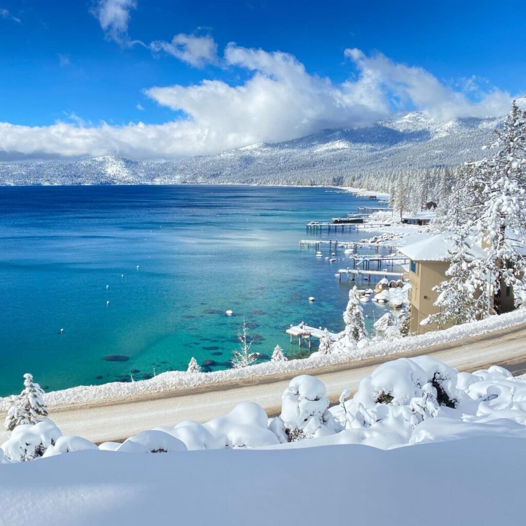 Blue lake surrounded by snow-covered mountains and pine trees under a sunny sky.